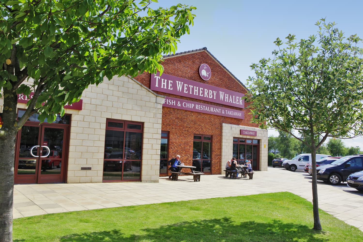 Red-brick fish and chip restaurant with outdoor seating, surrounded by green lawn and trees under a clear blue sky.