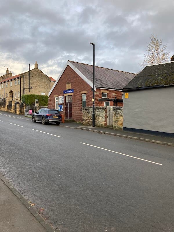Aberford Village Hall on a quiet street, with a brick exterior, parked car, and nearby stone buildings.