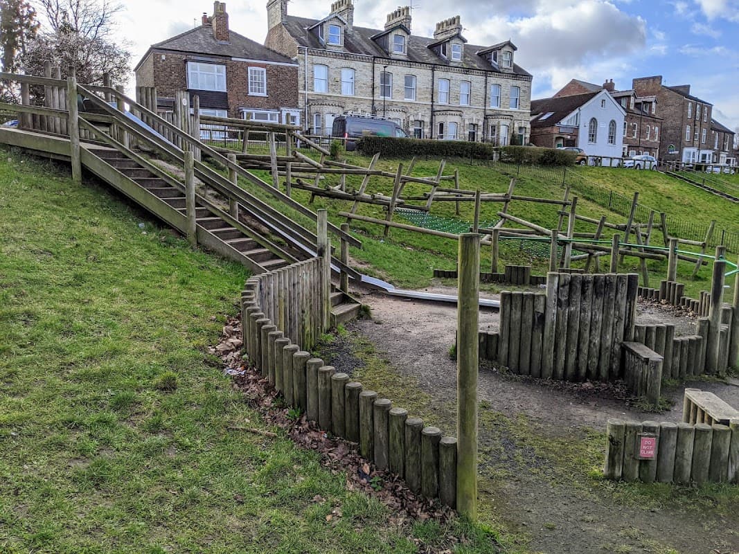 Wooden play structures and a grassy hill, with houses in the background under a partly cloudy sky.