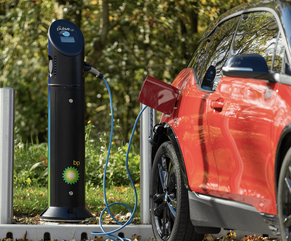 bp pulse charging station next to a red electric vehicle, with charging cable connected, set in a green outdoor environment.