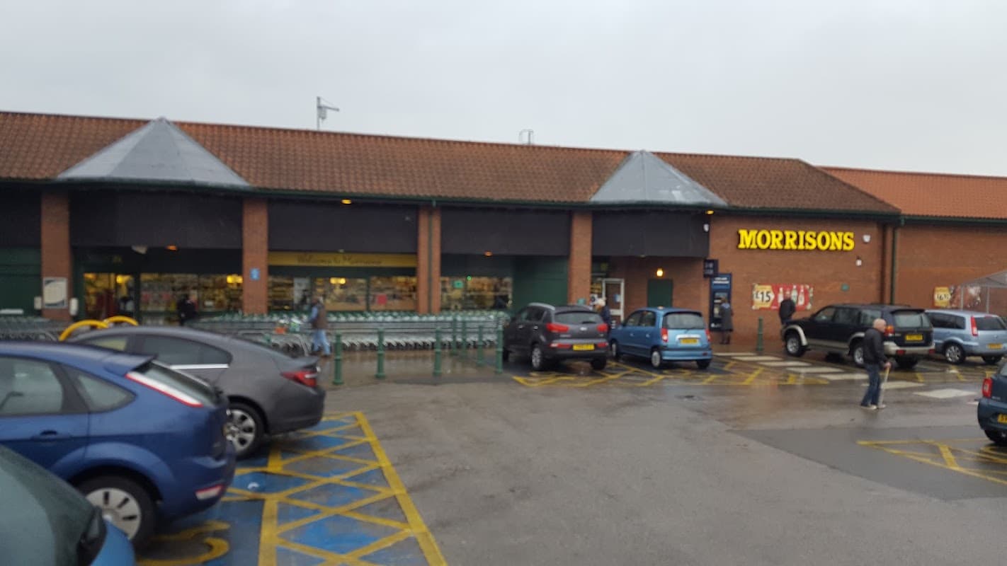 Morrisons supermarket in Acomb, Yorkshire, with cars parked in front and shopping carts outside.
