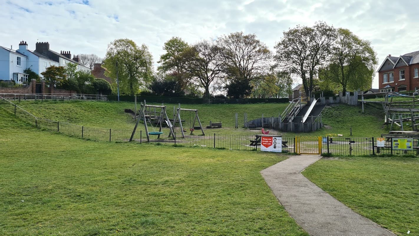 Play park with swings, slides, grassy areas, trees, and a pathway, surrounded by a fence in Acomb, Yorkshire.