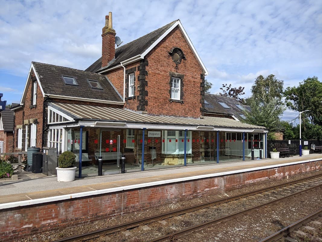 Brick building with large windows, covered platform, and train tracks under a blue sky at Poppleton Station Car Park.
