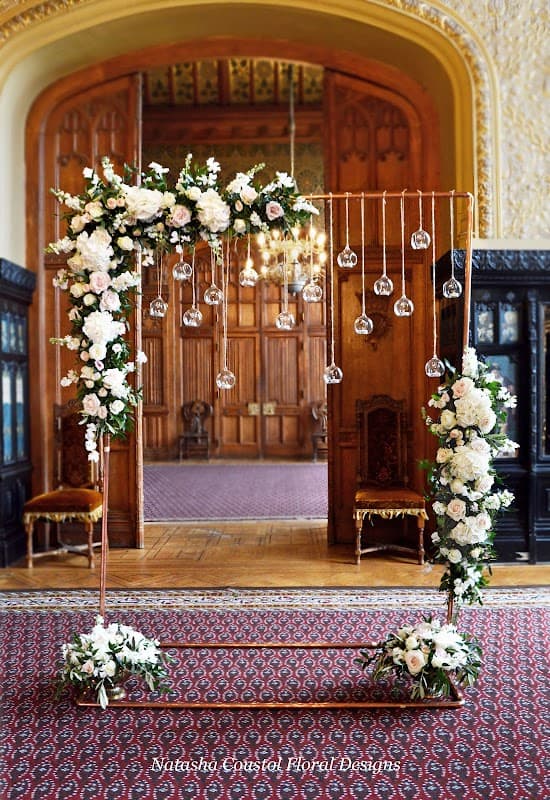 Floral arch adorned with white roses and hanging glass orbs, set in an elegant interior with wooden paneling.