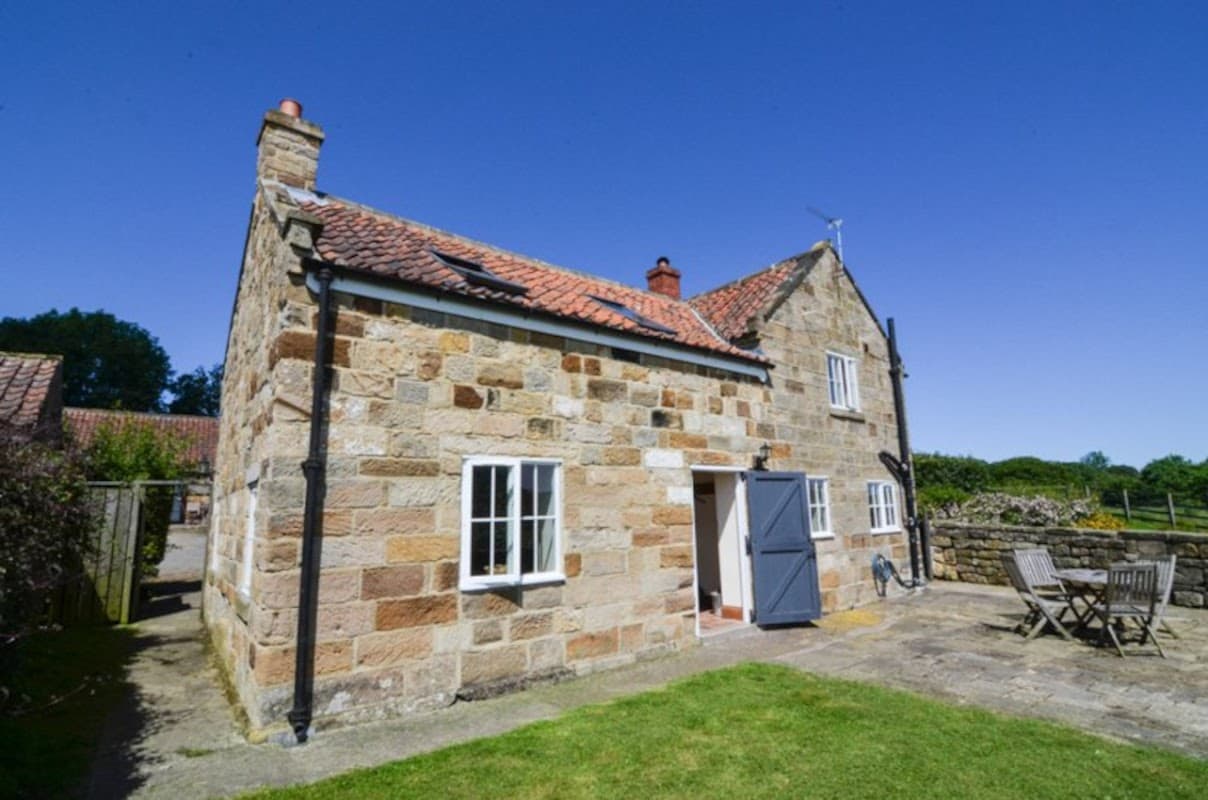 Stone cottage with a red-tiled roof, blue door, and outdoor seating area under a clear blue sky.
