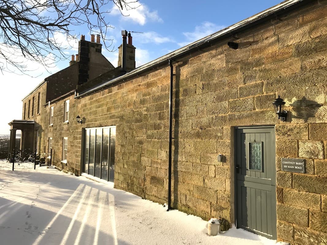 Chestnut Barn Holiday Cottage features a stone exterior, snow-covered ground, and large windows reflecting a sunny sky.