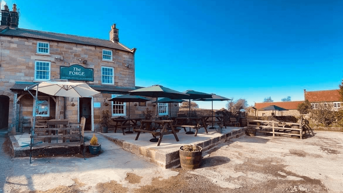 The Forge Inn features outdoor seating with umbrellas, stone exterior, and a clear blue sky in Aislaby, Yorkshire.
