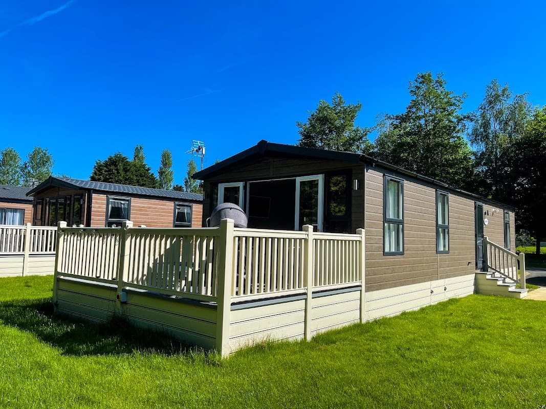 A modern lodge with a deck, surrounded by green grass and trees, under a clear blue sky in Allerthorpe, Yorkshire.