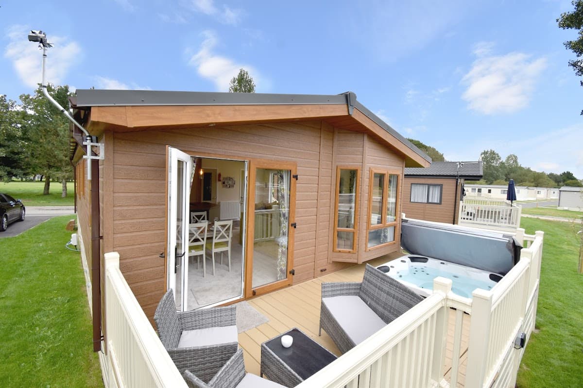 Modern wooden lodge with open doors, patio furniture, and a hot tub, surrounded by green grass and trees.