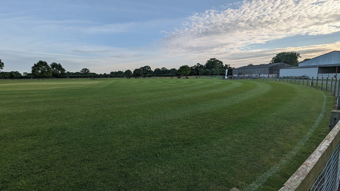 Lush green cricket field with neatly mowed stripes, surrounded by trees and a building in the background.