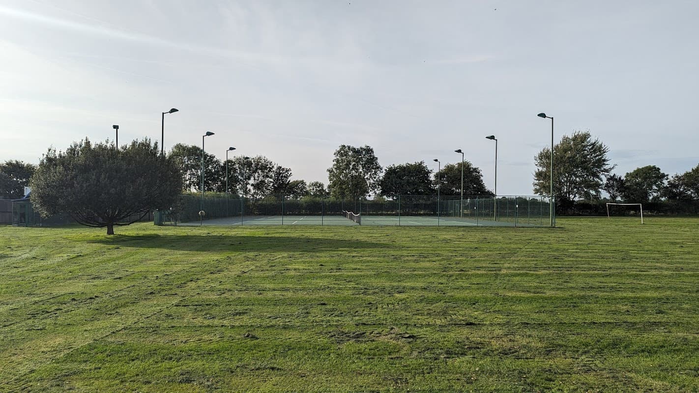 Tennis courts surrounded by neatly trimmed grass and trees, with goalposts in the background under a clear sky.