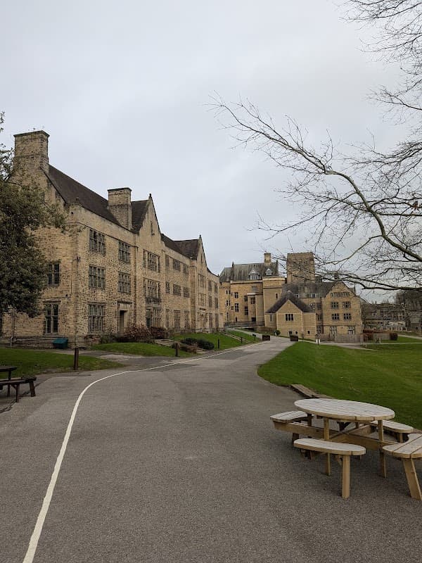 Stone buildings with large windows line a pathway, surrounded by grass and trees, under a cloudy sky.