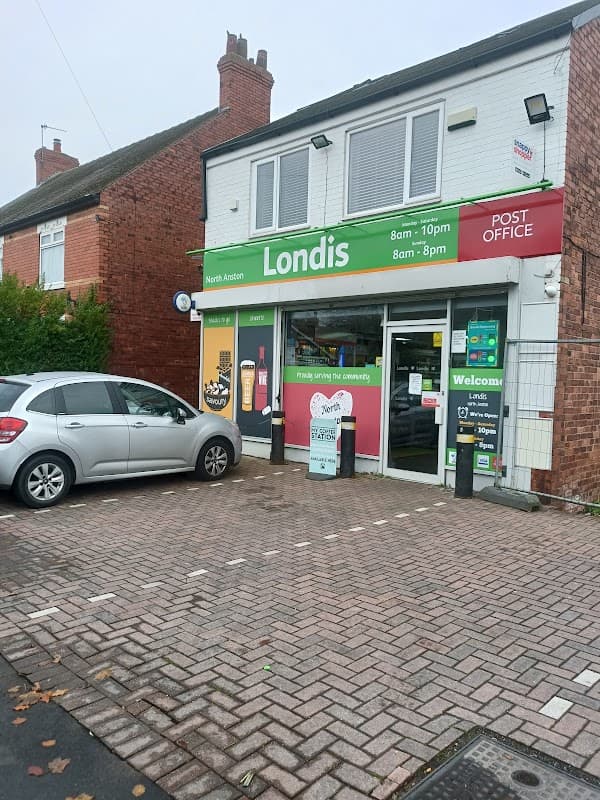 Londis storefront with a Post Office sign, open hours displayed, and a gray car parked in front on a brick pavement.