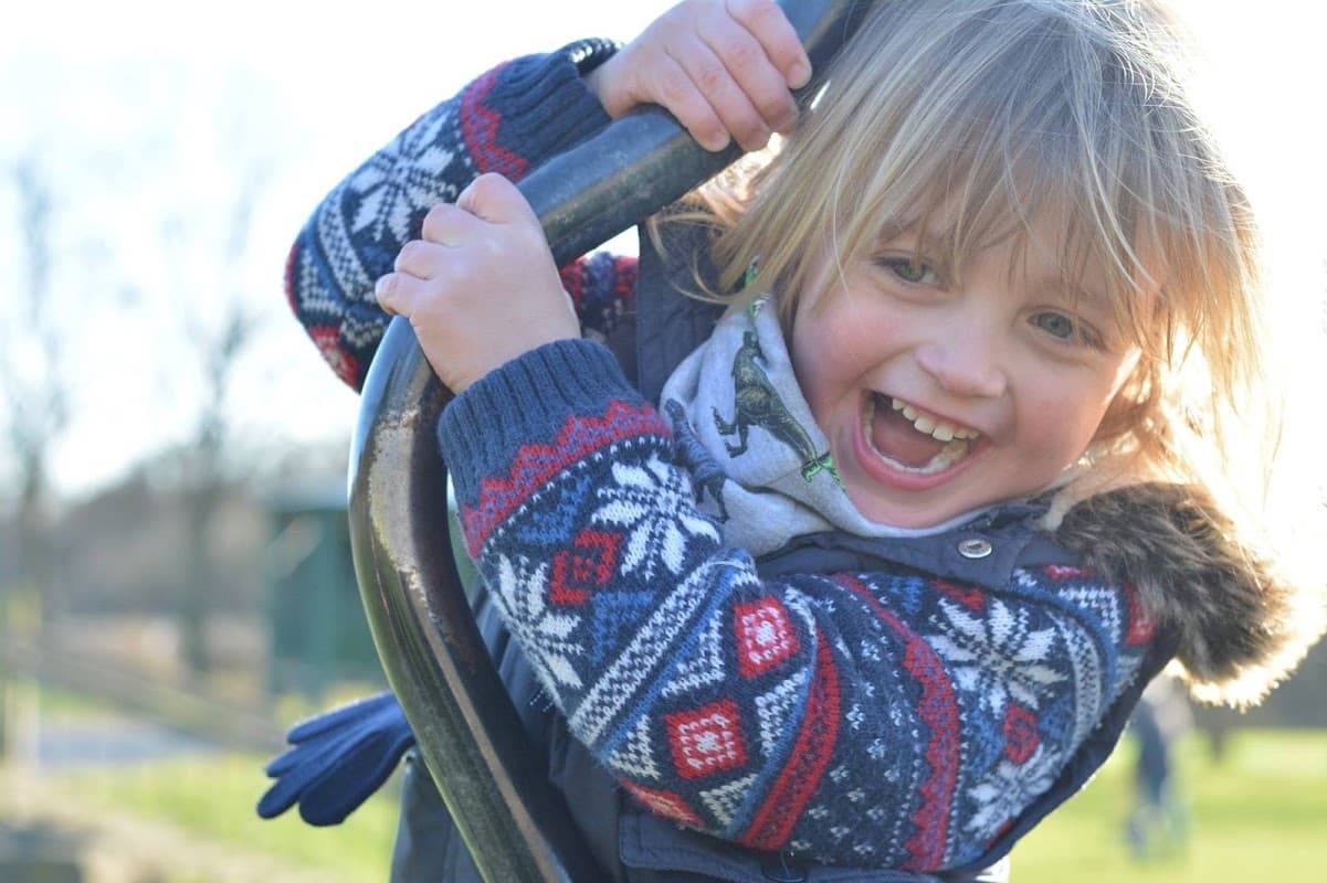 A young child with blonde hair smiles joyfully while climbing on a playground structure, wearing a colorful sweater.
