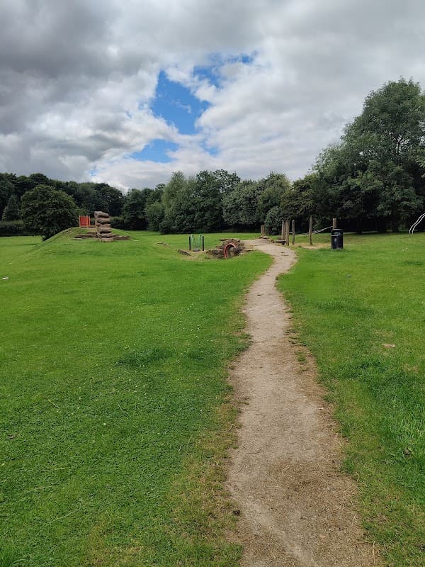Pathway through a grassy area with trees, play equipment, and a cloudy sky in Kendal Park, Anston.