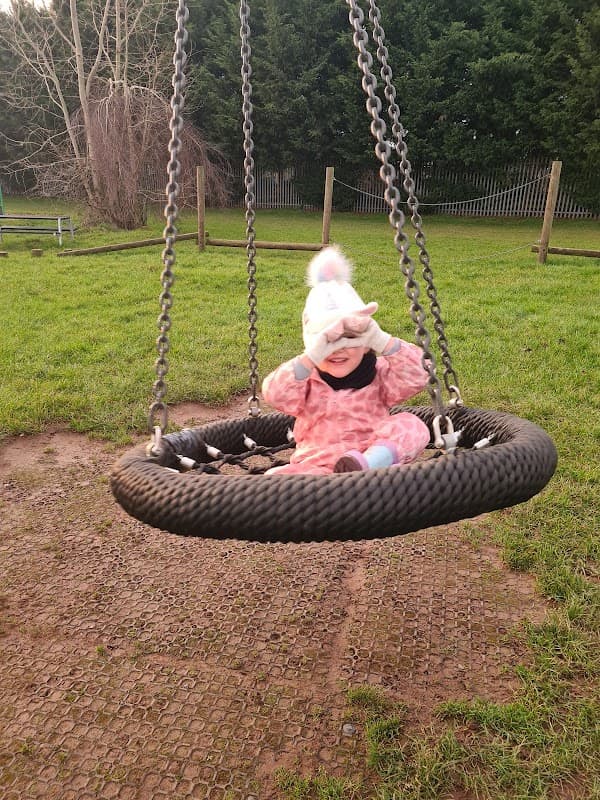 A child in a pink outfit sits on a large swing, smiling and shielding their eyes from the sun in a grassy park.
