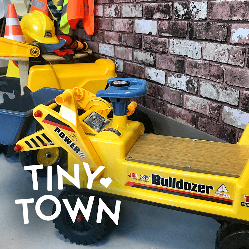 Bright yellow toy bulldozer with a wooden seat, construction helmets, and safety cones against a brick wall backdrop.