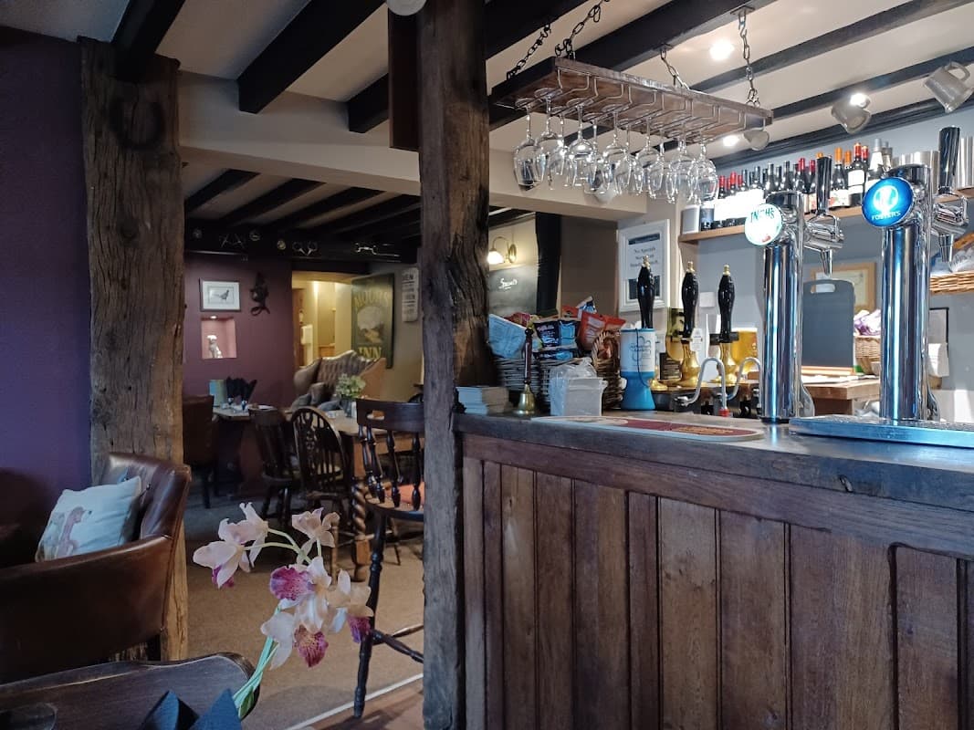 Cozy bar interior with wooden beams, a counter, hanging glassware, and seating areas in a rustic setting.