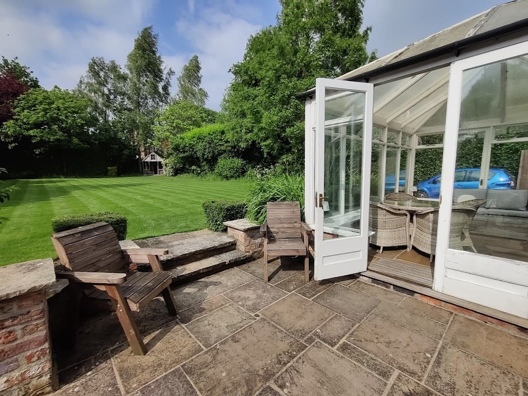 Sunlit patio area with wooden chairs, a glass conservatory, and a lush green garden in Appleton Roebuck, Yorkshire.