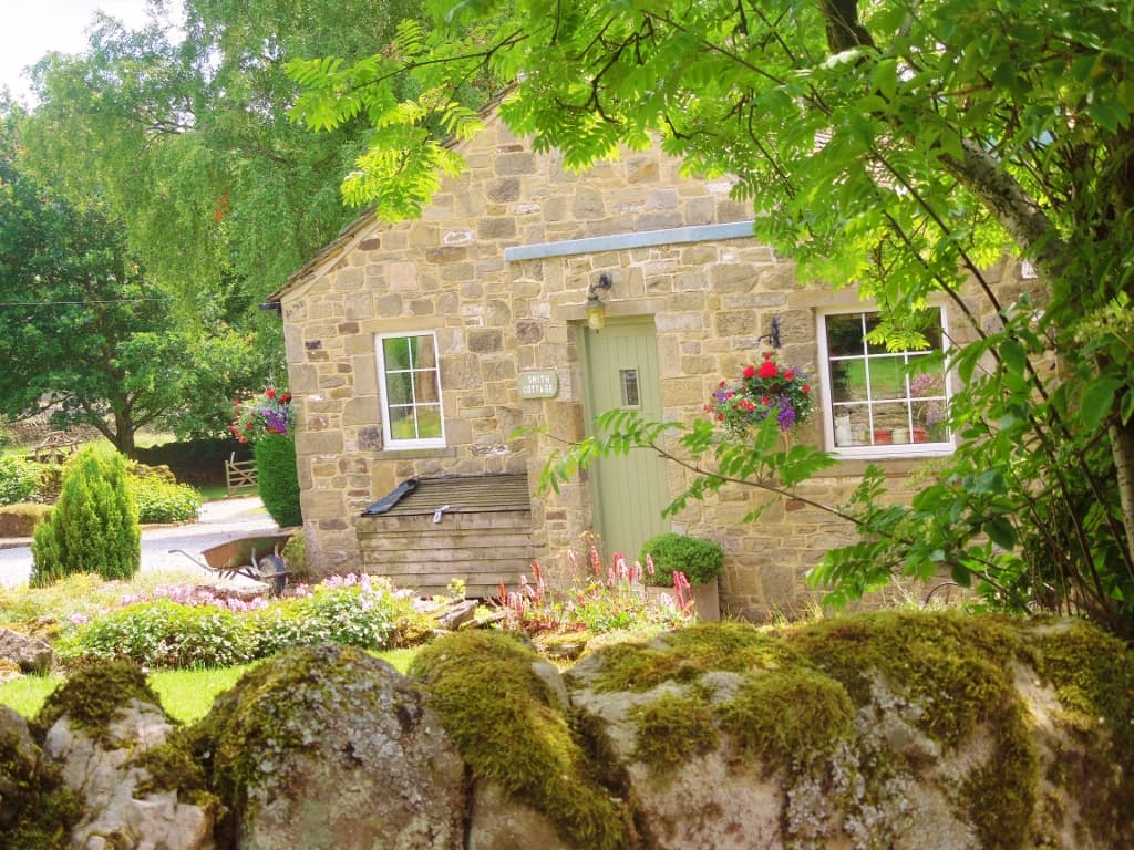 Charming stone cottage with green door, flower boxes, and lush garden, set against a backdrop of trees and a mossy wall.