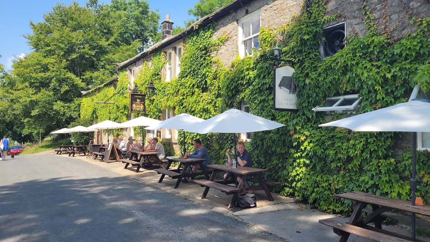 Quaint pub with outdoor seating under umbrellas, surrounded by lush greenery and stone walls in a sunny Yorkshire setting.