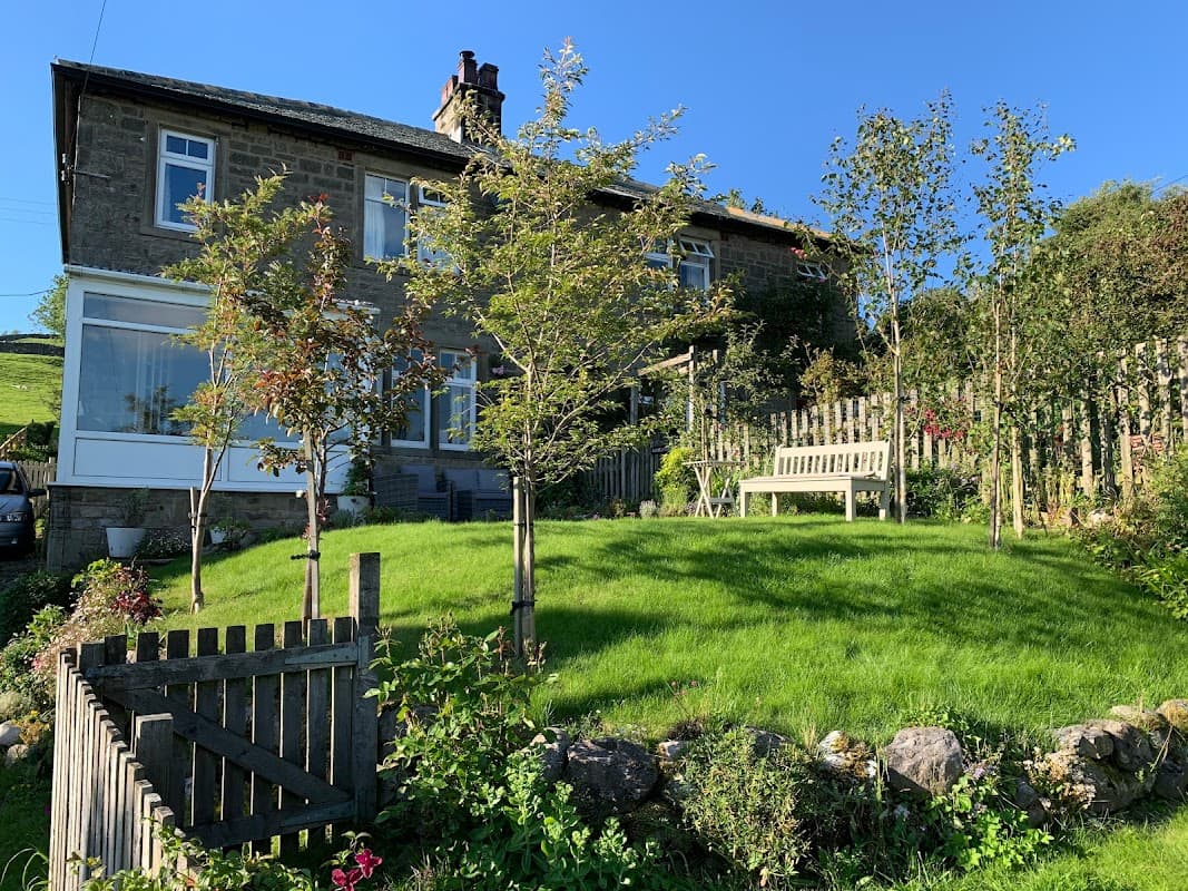 Fellside Cottage with a lush green garden, trees, and a wooden bench, set against a clear blue sky.