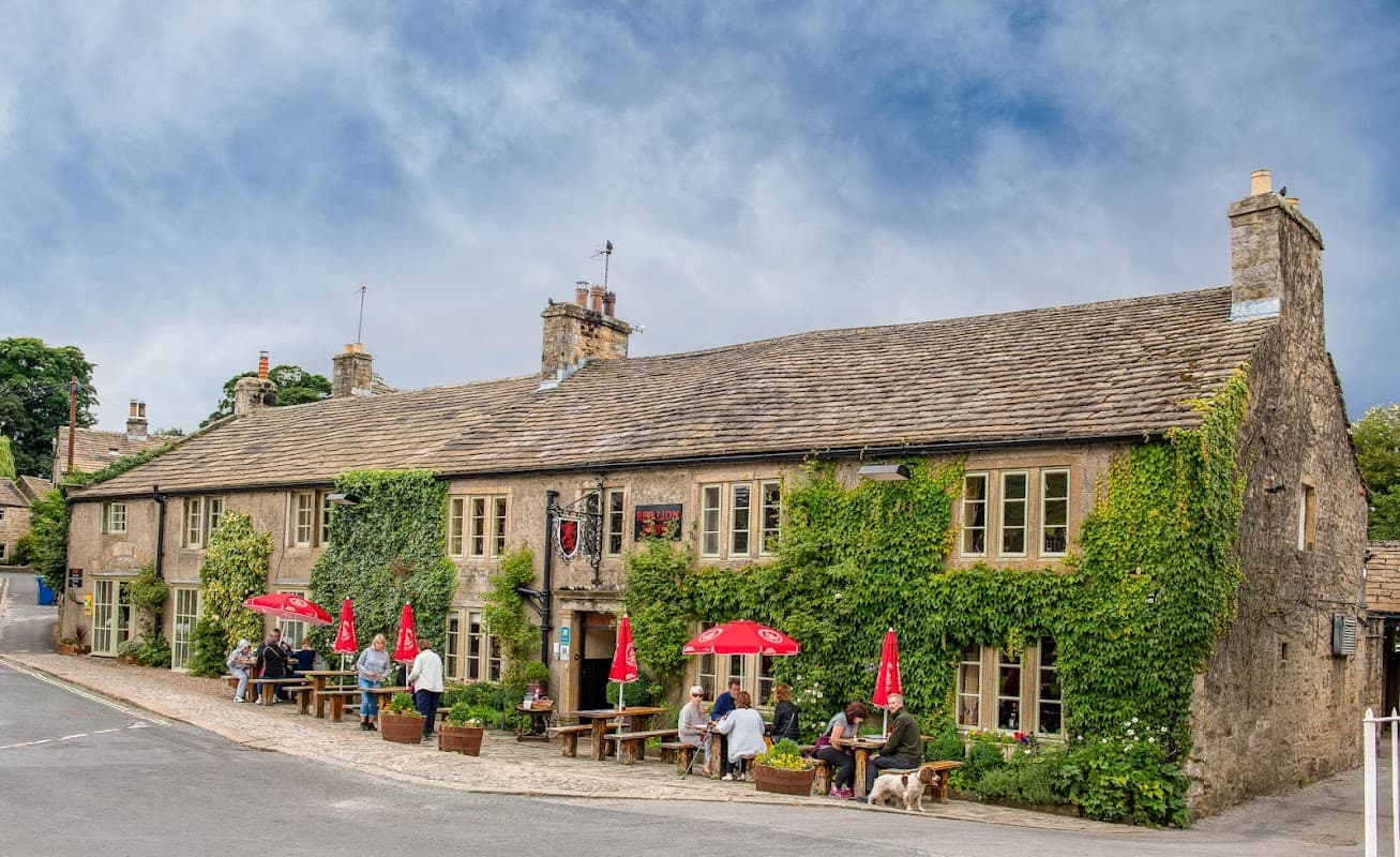 Historic stone pub with outdoor seating, red umbrellas, and patrons enjoying the scenic Yorkshire village.