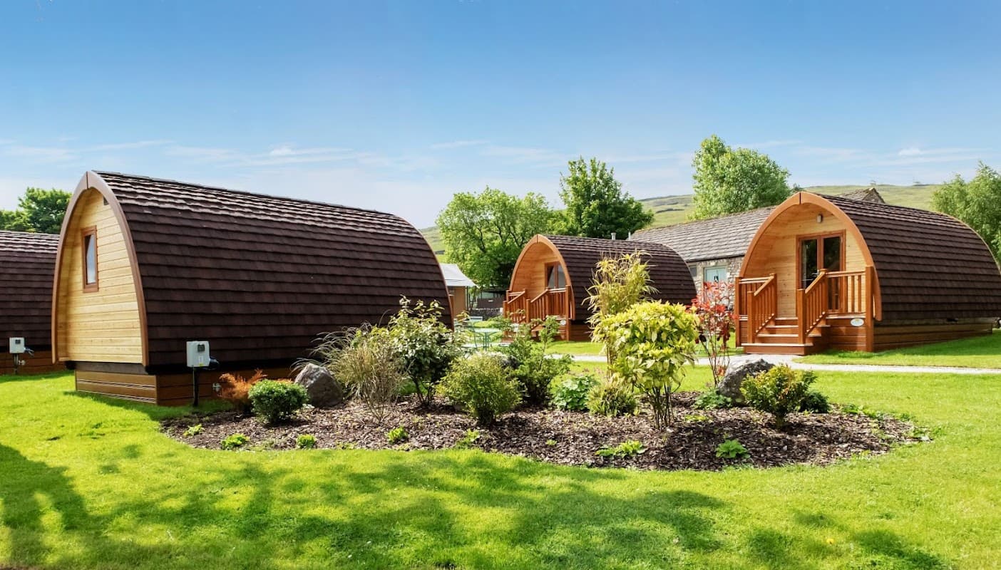 Wooden holiday lodges with arched roofs surrounded by lush green grass and colorful shrubs under a clear blue sky.