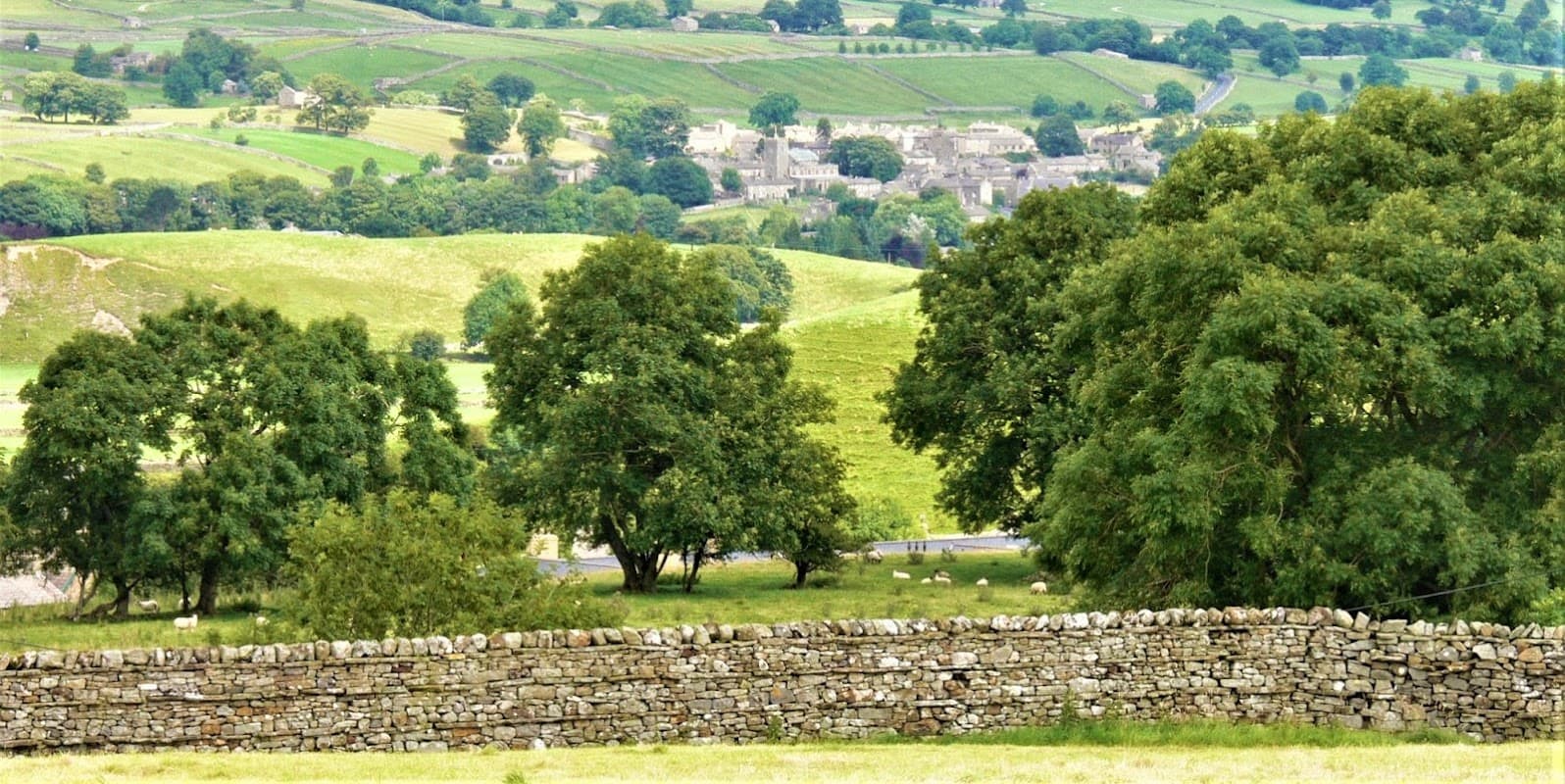 View of lush green hills, trees, and a stone wall in Askrigg, Yorkshire, with a distant village in the background.