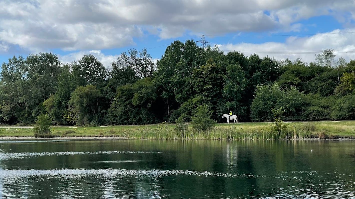 A serene lake reflects the sky, with lush greenery and a person on horseback in the distance along a path.