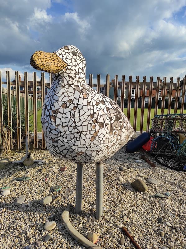 Mosaic seagull sculpture stands on a pebbled area, with a grassy field and wooden fence in the background.