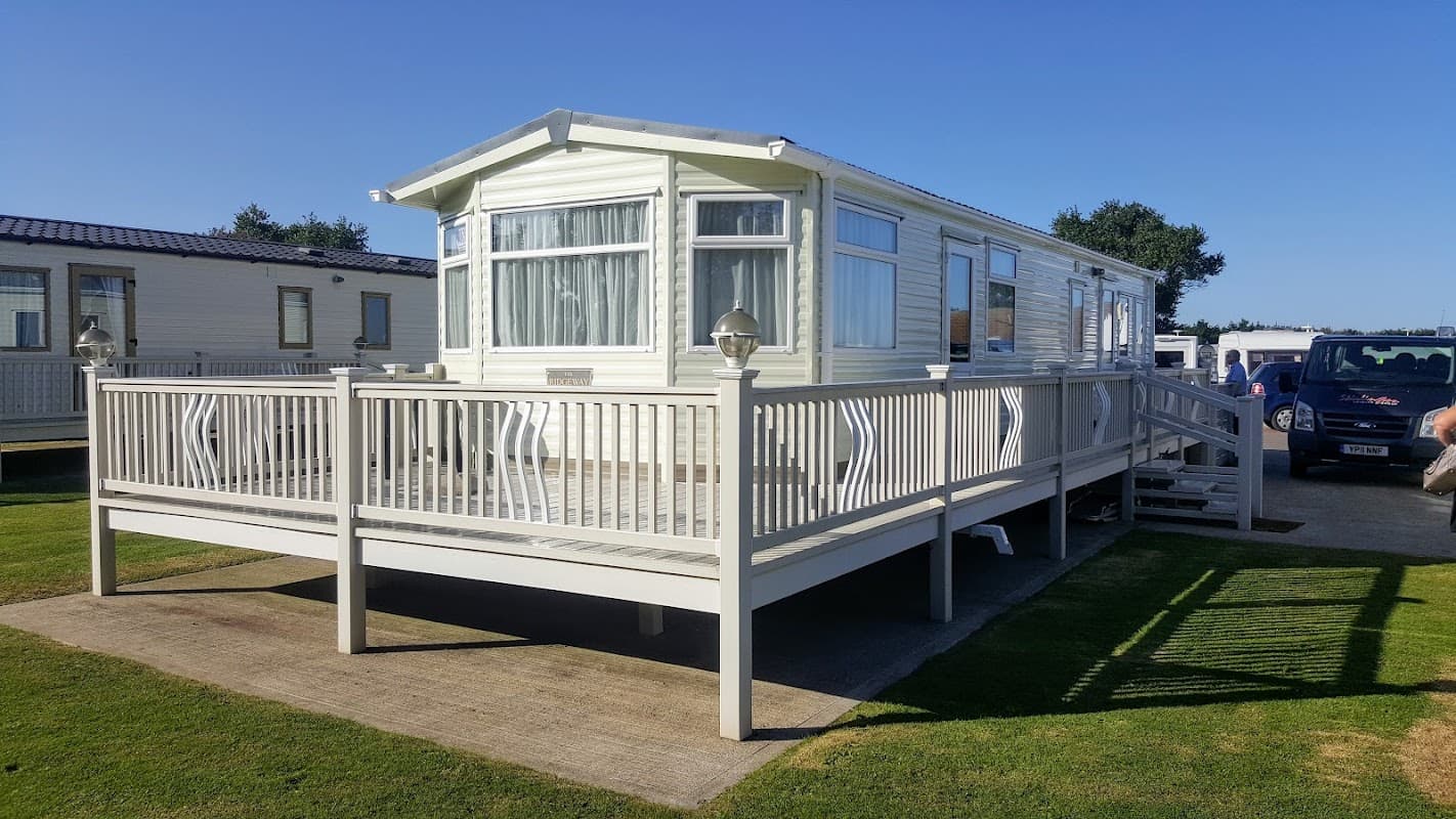 A holiday home with a deck, surrounded by green grass and parked vehicles under a clear blue sky at Skirlington Leisure Park.