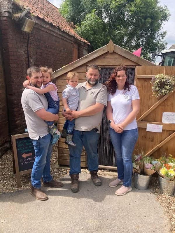 A family stands in front of a wooden shed at The Egg Shed Farm Shop, with flowers and a chalkboard sign nearby.