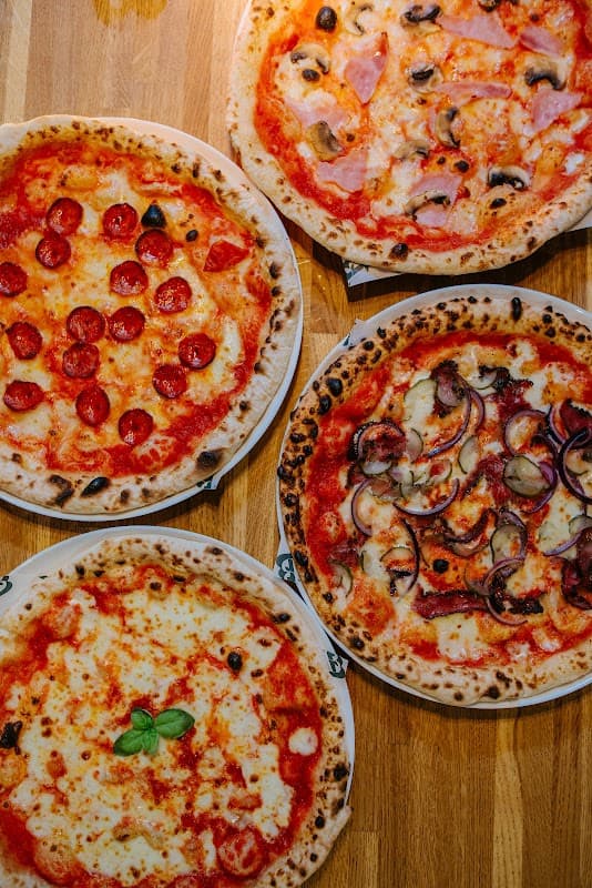 Four pizzas on wooden table: pepperoni, ham and mushroom, margherita with basil, and a cheesy pizza with red onions.