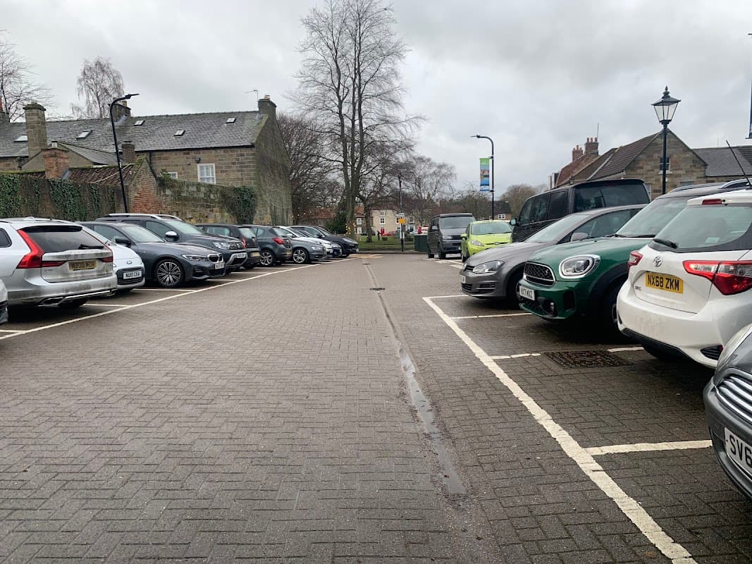 Car park with various parked vehicles, trees in the background, and a cloudy sky in Ayton, Yorkshire.