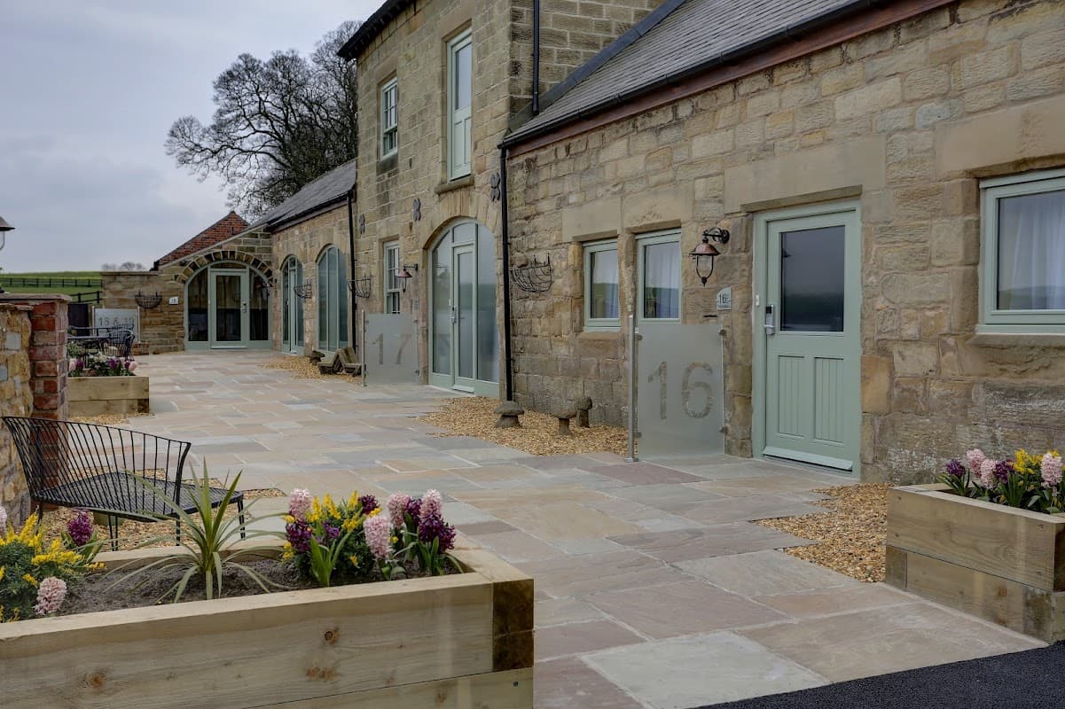 Historic stone building with large windows, numbered doors, and landscaped garden beds featuring flowers and seating.