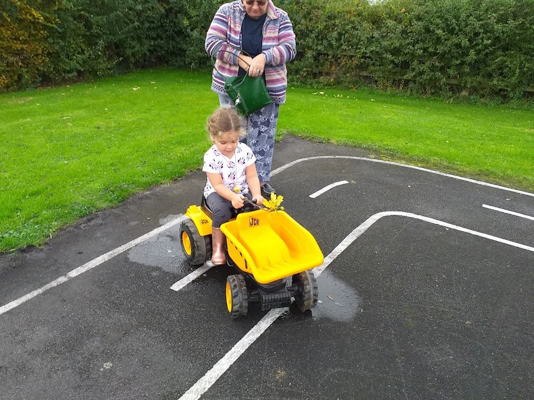 A child rides a yellow toy tractor on a play area path while an adult watches nearby in a green field.