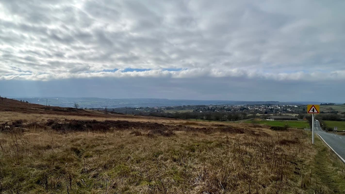 Scenic view from Baildon Moor Parking, featuring rolling hills, distant village, and cloudy sky.