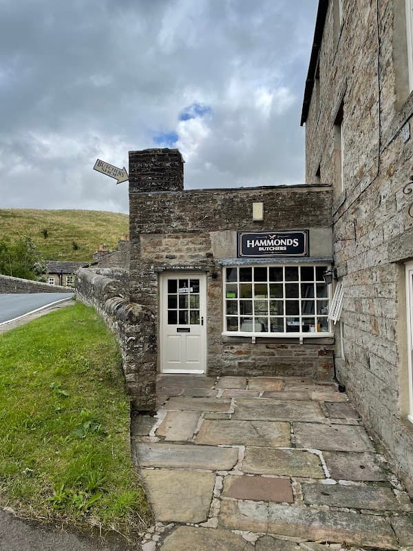 Hammonds Butchers shop with stone facade, large window, and sign, set against a green hillside in Bainbridge, Yorkshire.