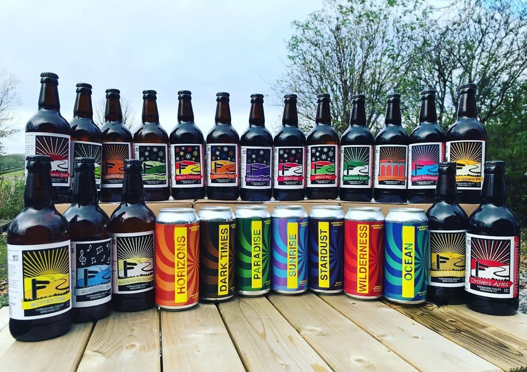 A display of various beer bottles and cans from Yorkshire Dales Brewery, featuring colorful labels and scenic outdoor backdrop.