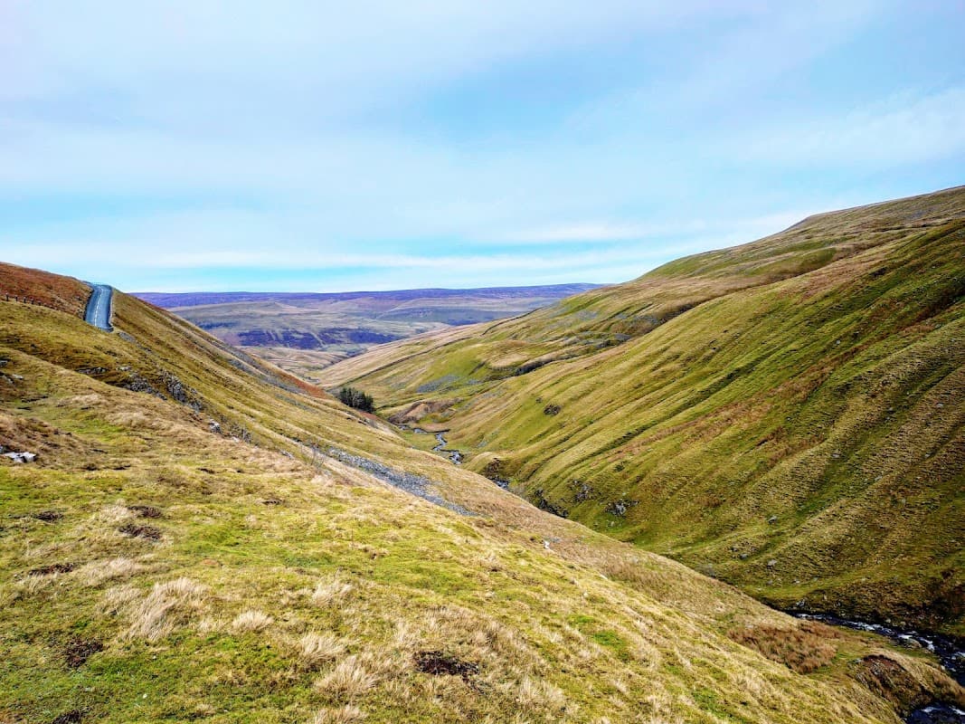Rolling green hills and a winding road through the Yorkshire Dales, with a river flowing in the valley below.