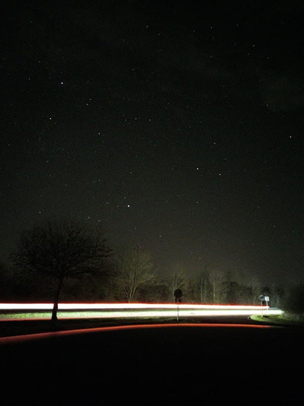 Starry night sky over a dark road with light trails from passing vehicles and silhouetted trees.