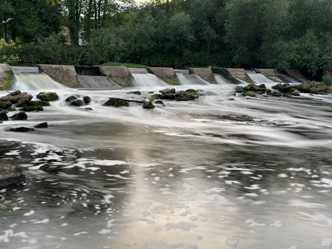 Serene water flowing over a weir, surrounded by lush greenery and smooth stones along the riverbank.