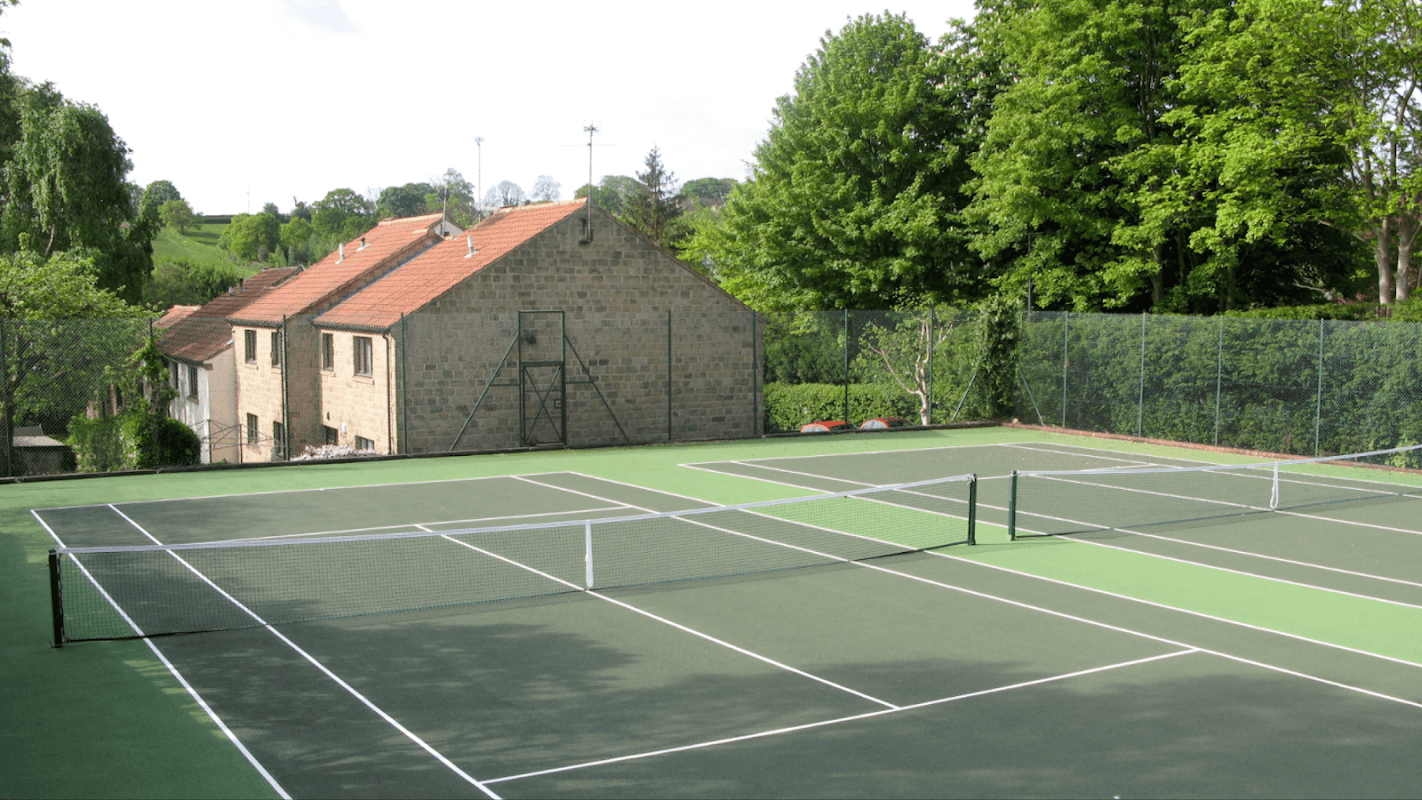 Tennis courts with green surfaces and white lines, surrounded by trees and a stone building in Bardsey, Yorkshire.