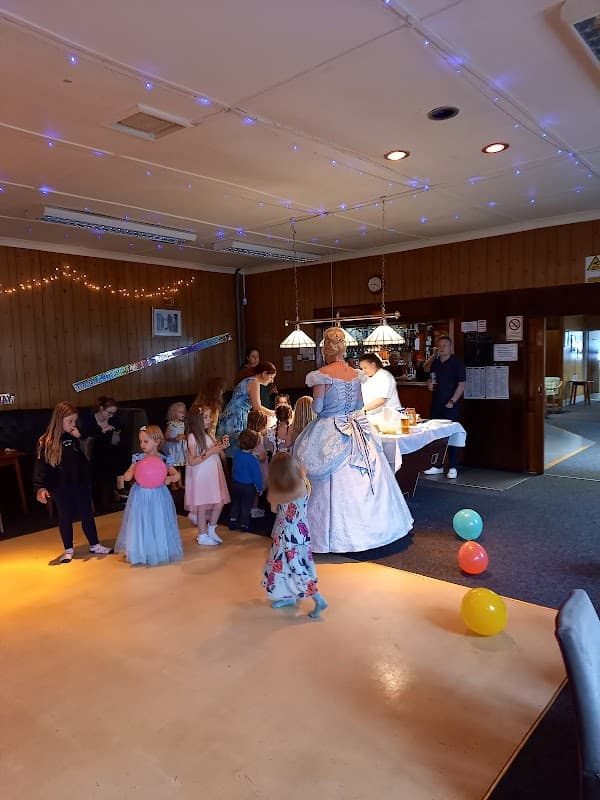 A festive event at Barlow Village Club with children dancing, balloons, and a woman in a blue gown interacting with guests.