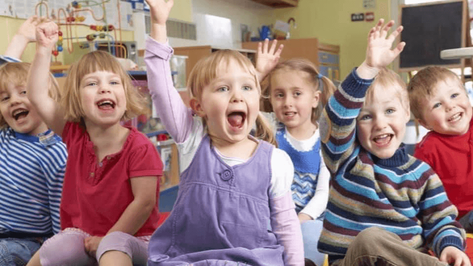 Children sitting in a classroom, enthusiastically raising their hands, smiling and engaging with each other.