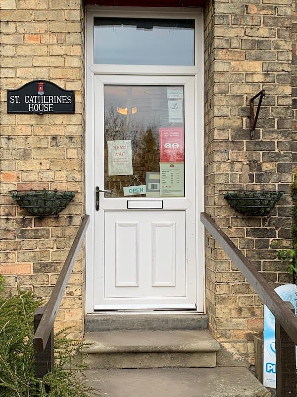 White door with glass panel, "St Catherine's House" sign, and potted plants on either side. Open sign visible.