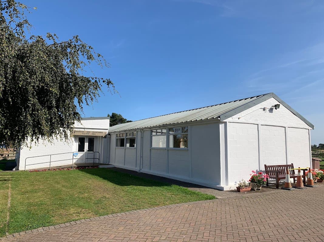 White village hall with a sloped roof, surrounded by green grass and a bench, under a clear blue sky.