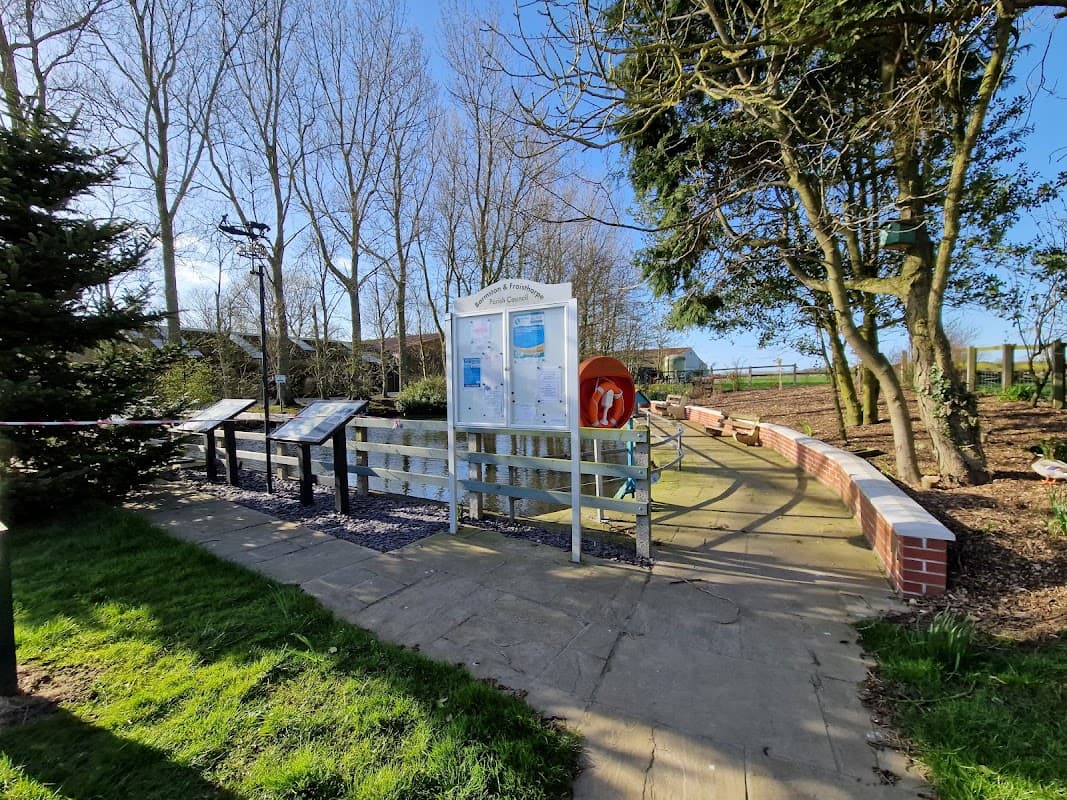 Barmston village pond with informational boards, a lifebuoy, trees, and a path surrounded by green grass.