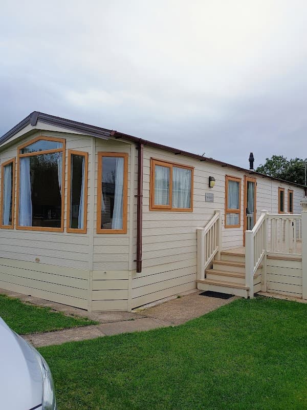 A holiday park cabin with large windows, a wooden deck, and green grass in Barmston, Yorkshire, under a cloudy sky.
